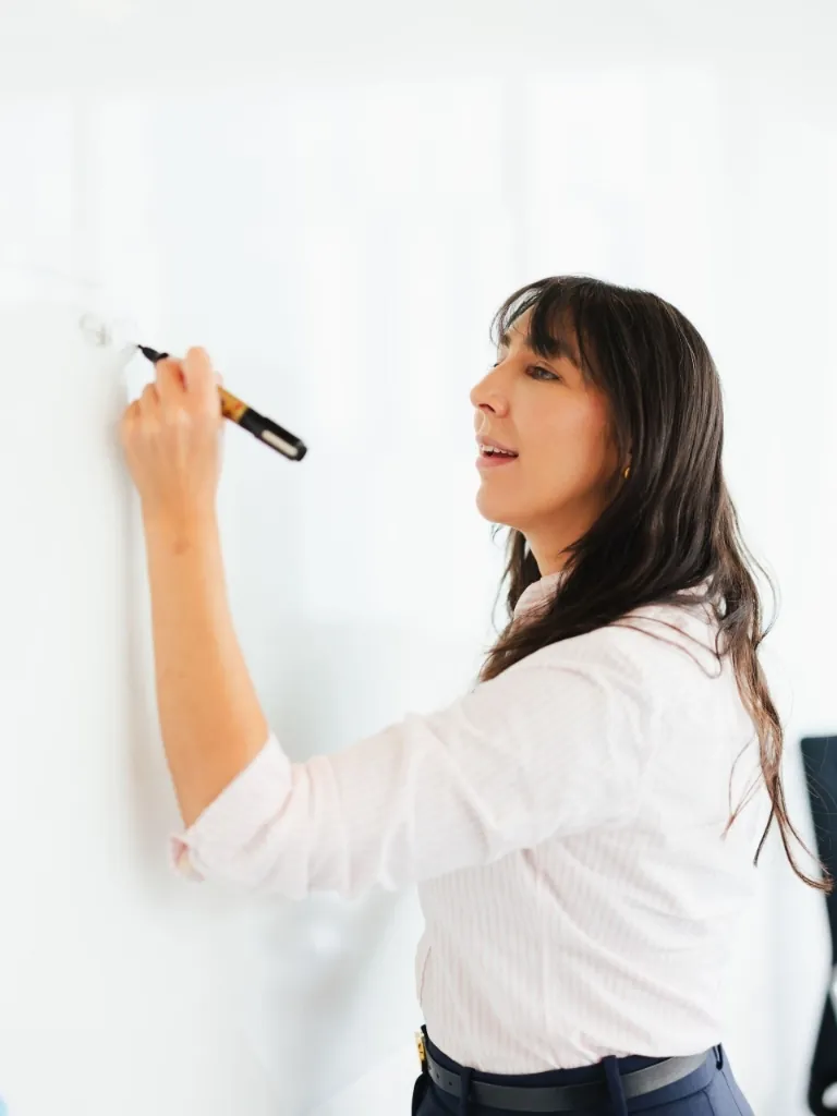 A professional woman with dark hair writing strategic notes on a whiteboard during a planning session.