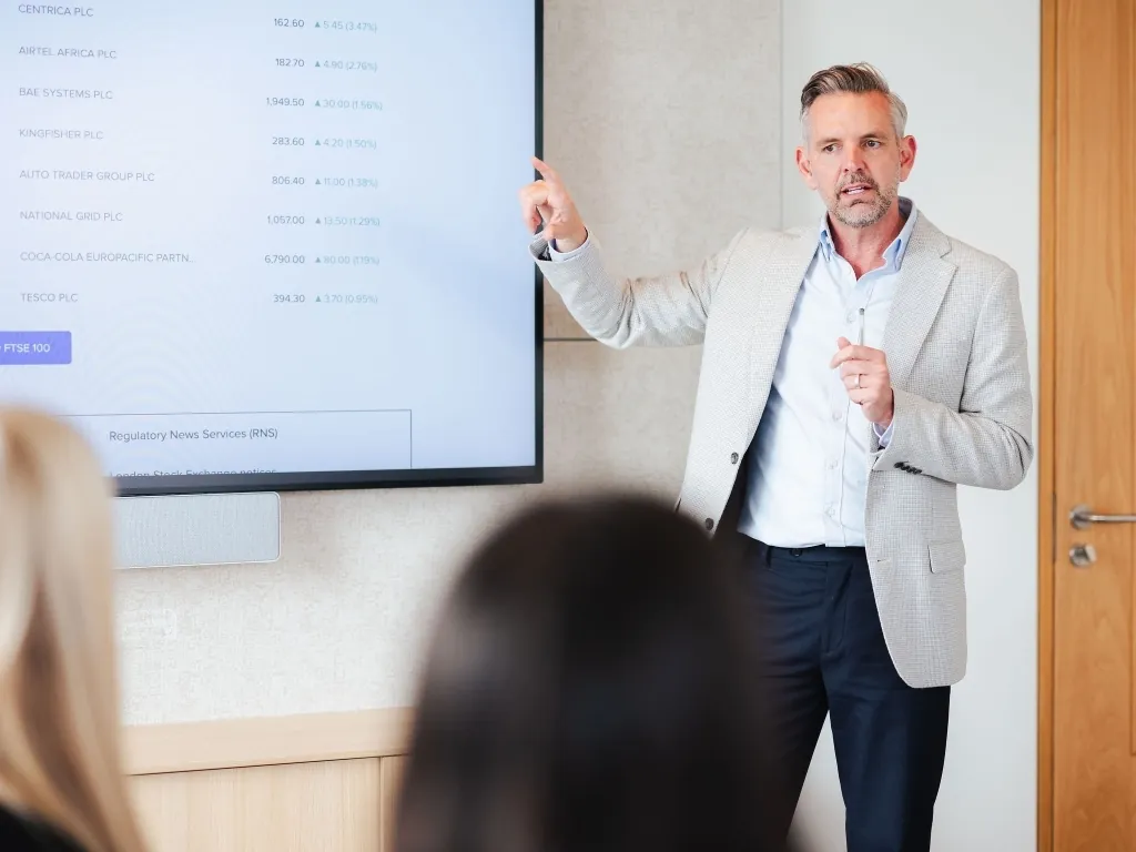 A male professional in a light grey blazer pointing to a screen displaying financial data and stock indices.
