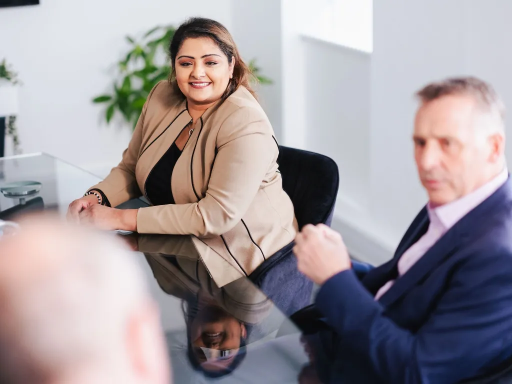 A professional woman in a tan blazer smiling during a strategic board meeting at a glass table.