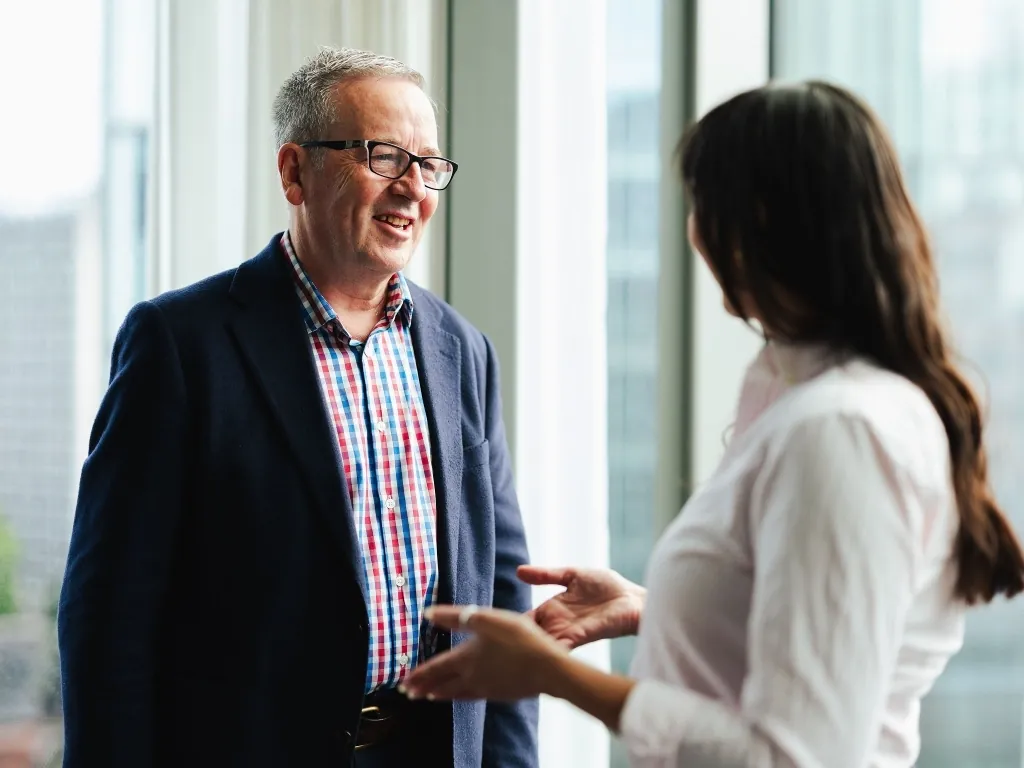 A smiling man in a blazer and checkered shirt engaged in a professional conversation with a colleague.