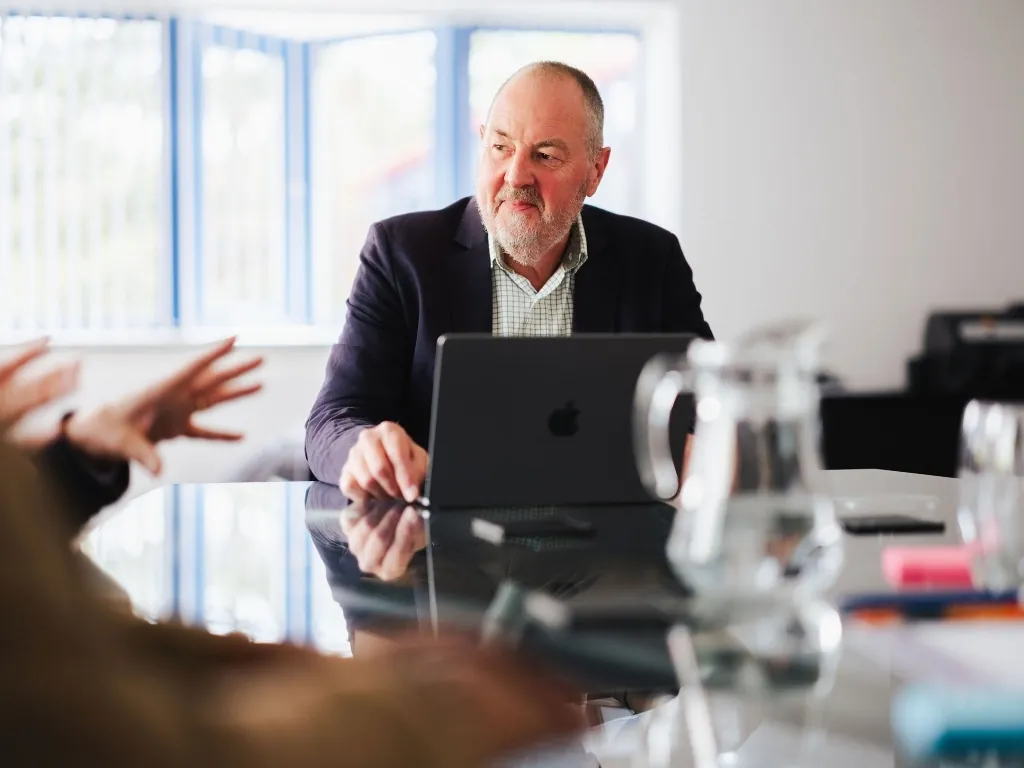 A senior business consultant with a beard working on a laptop during a collaborative office meeting.