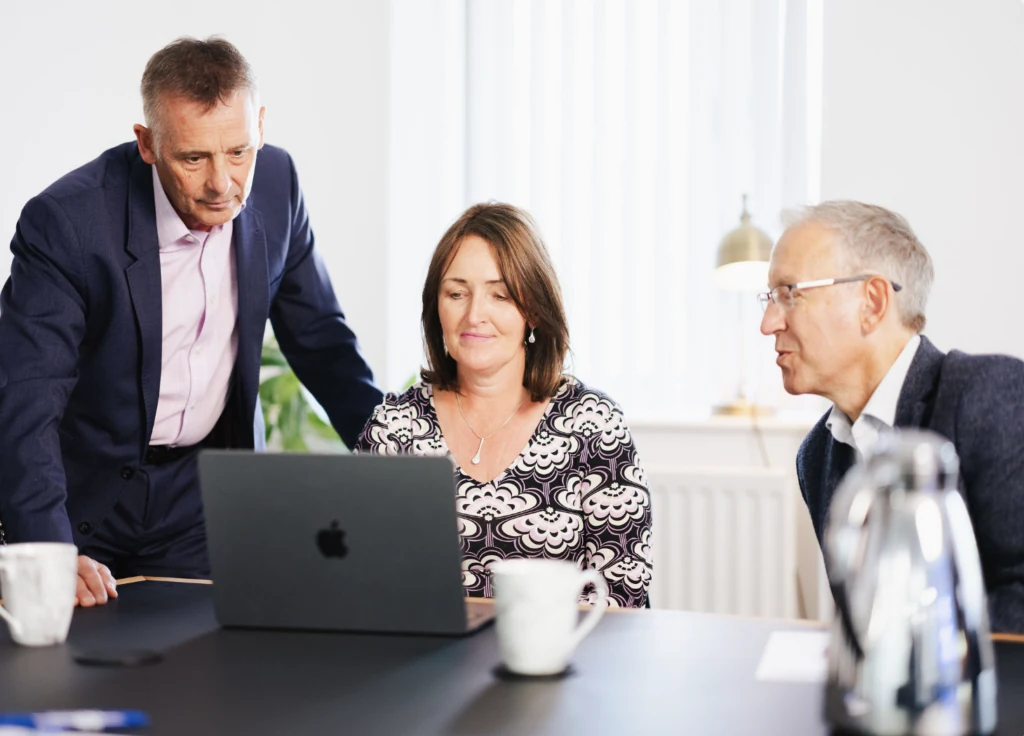 3 people looking at a laptop computer 2 sitting down and 1 standing with a hand on the desk