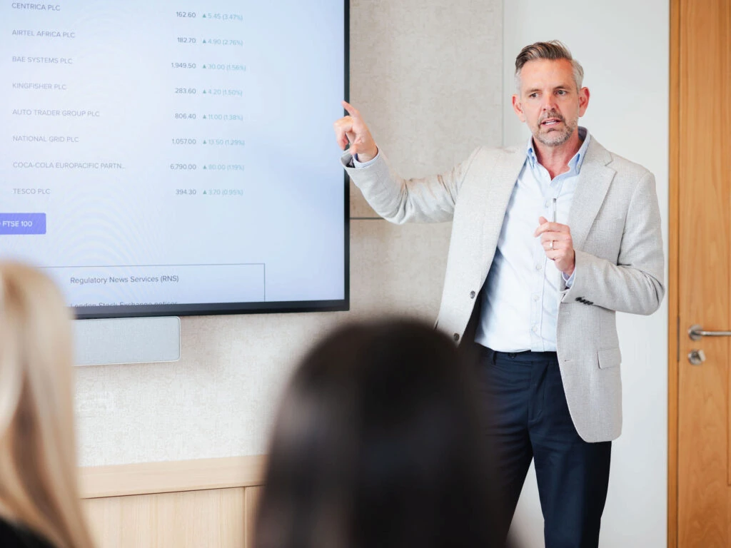 a man standing by a large screen showing financial data with 2 people watching in the foreground